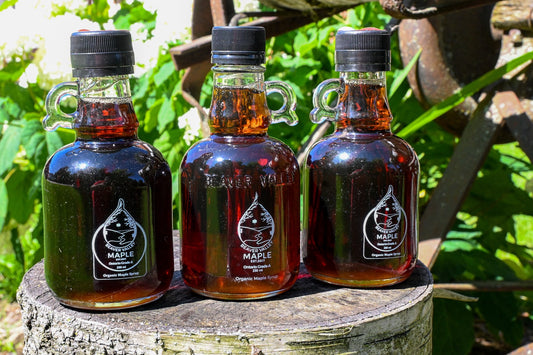 Three bottles of Beaver Valley Organic Maple Syrup on a weathered stump with rustic farm equipment and green foliage