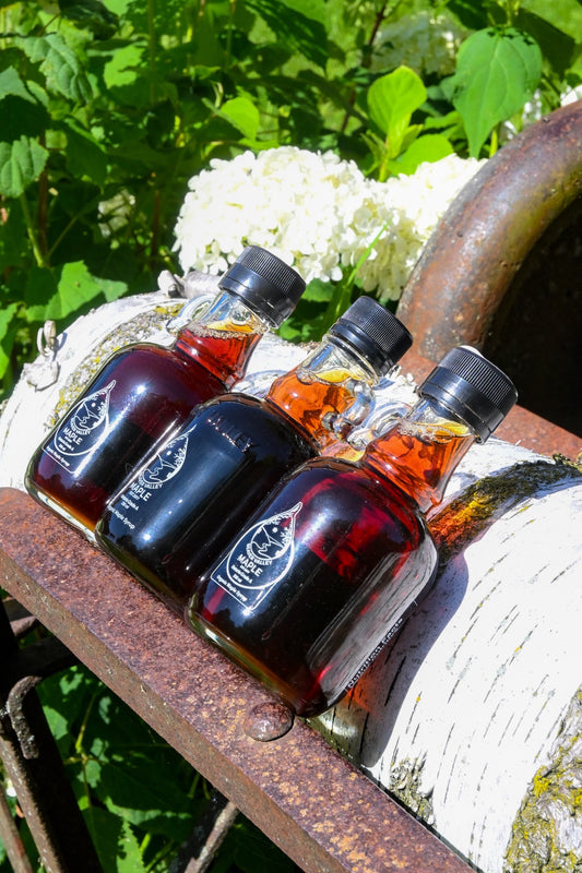 Three bottles of Beaver Valley Maple Syrup displayed on rustic farm equipment with white flowers and greenery in the background